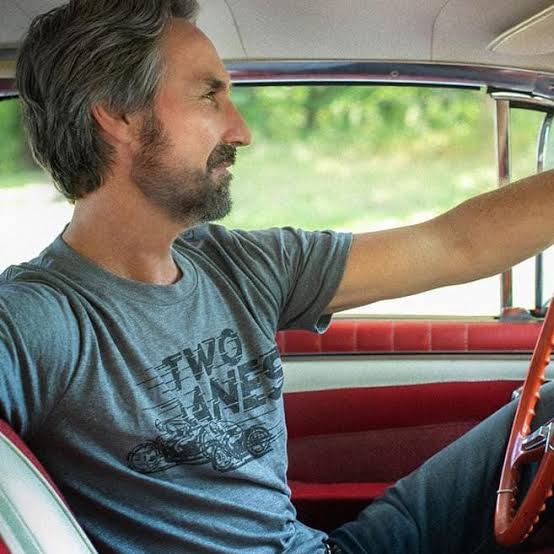 Mike Wolfe driving a vintage car in a Two Lanes tee, dashboard and steering wheel visible