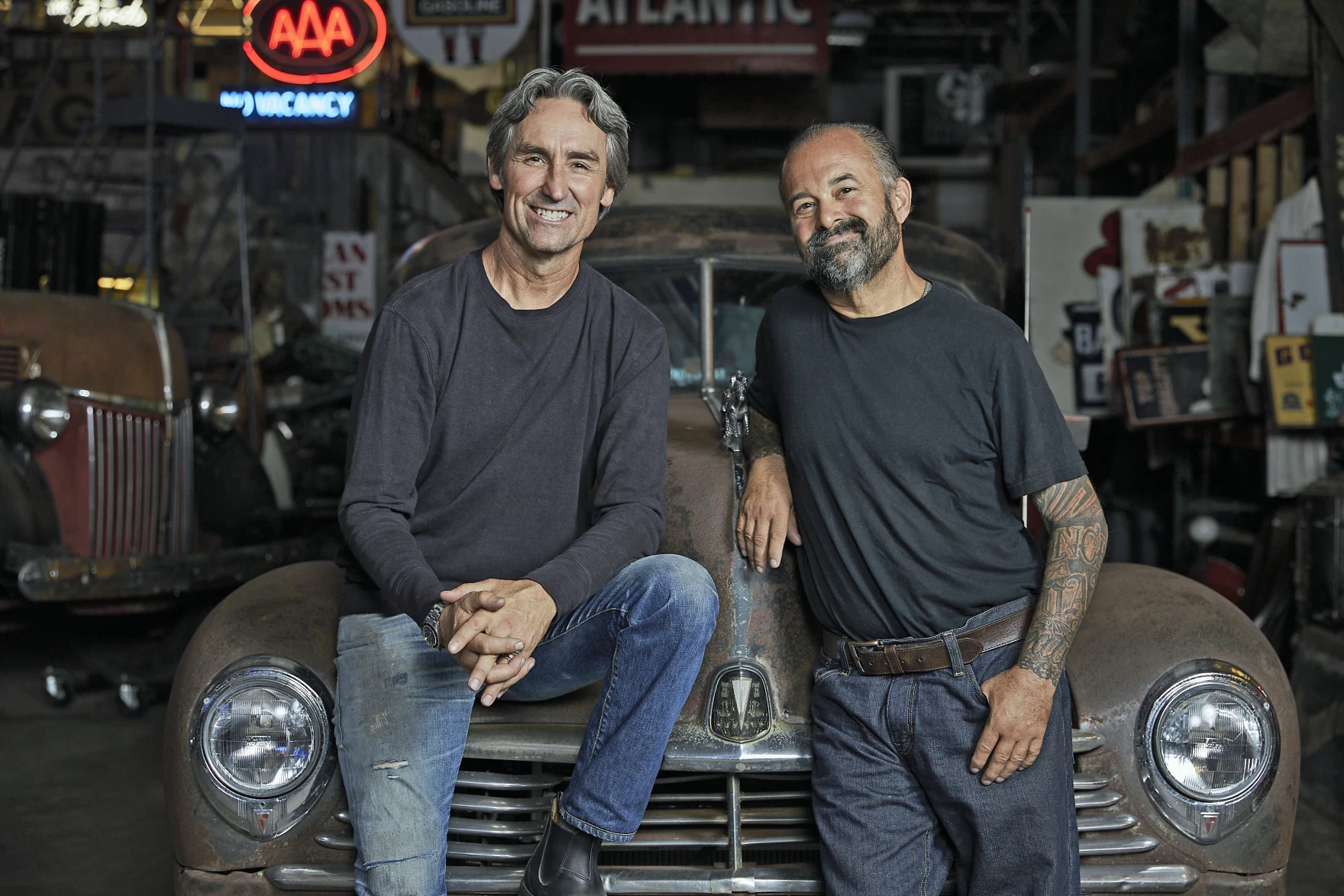 Mike Wolfe and Frank Fritz smiling together inside the Antique Archaeology shop, leaning on a vintage car hood surrounded by neon signs and collected memorabilia